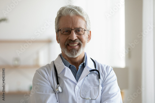 Head shot portrait of smiling older male general practitioner in eyeglasses and medical coat posing in clinic. Happy sincere trusted middle aged doctor looking at camera, medical service concept.