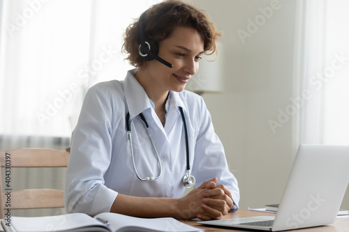 Pleasant happy young female doctor physician wearing headset with microphone, looking at laptop screen, giving medical advices or professional healthcare consultation to patient distantly online.