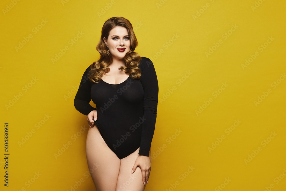 Beautiful plus-size model woman with bright makeup in black bodysuit posing over yellow background, isolated. Young sexy plump woman Beautiful plus-size model woman with bright makeup in black bodysuit posing over yellow background, isolated. Young sexy plump woman