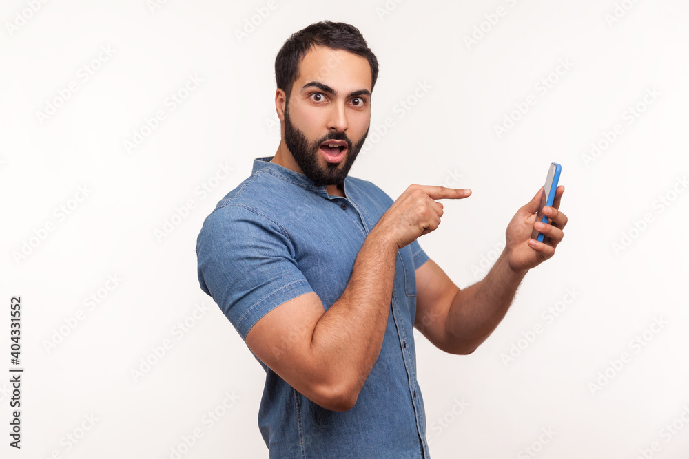 Side view surprised man pointing finger at smartphone display, shocked with bonuses, easy booking, online applications. Indoor studio shot isolated on white background Side view surprised man pointing finger at smartphone display, shocked with bonuses, easy booking, online applications. Indoor studio shot isolated on white background