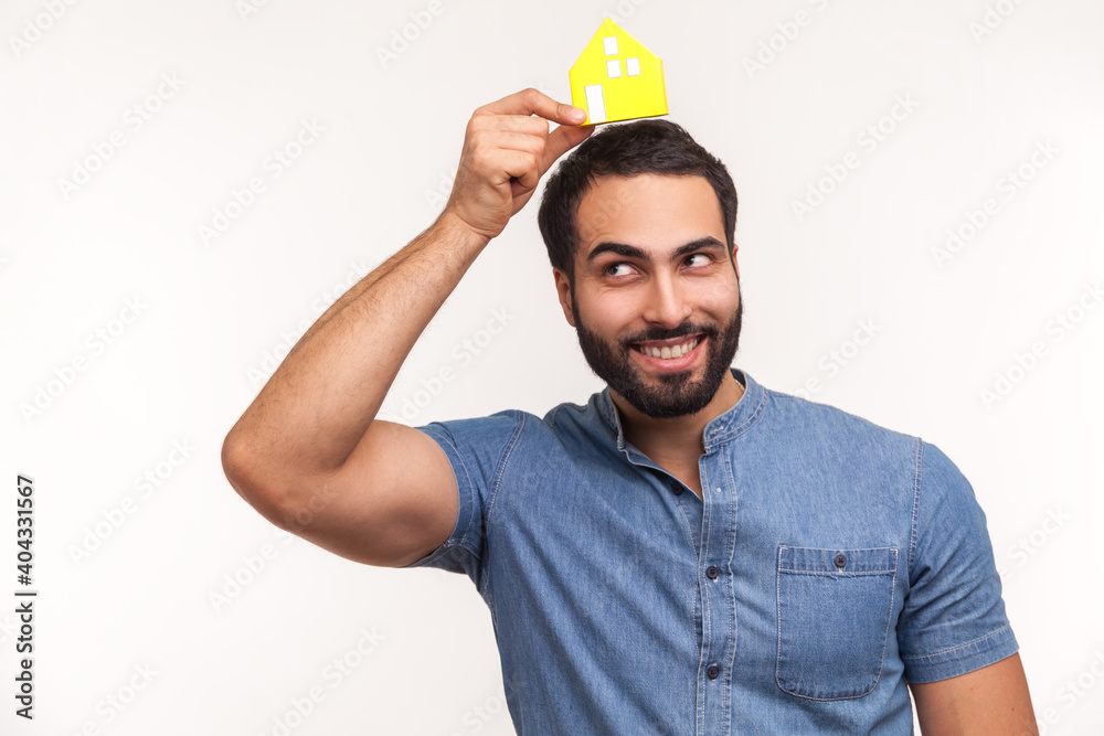 Naklejka premium Happy dreamy man with beard holding little paper house on head, imagining his own apartment, housing purchase, real estate. Indoor studio shot isolated on white background