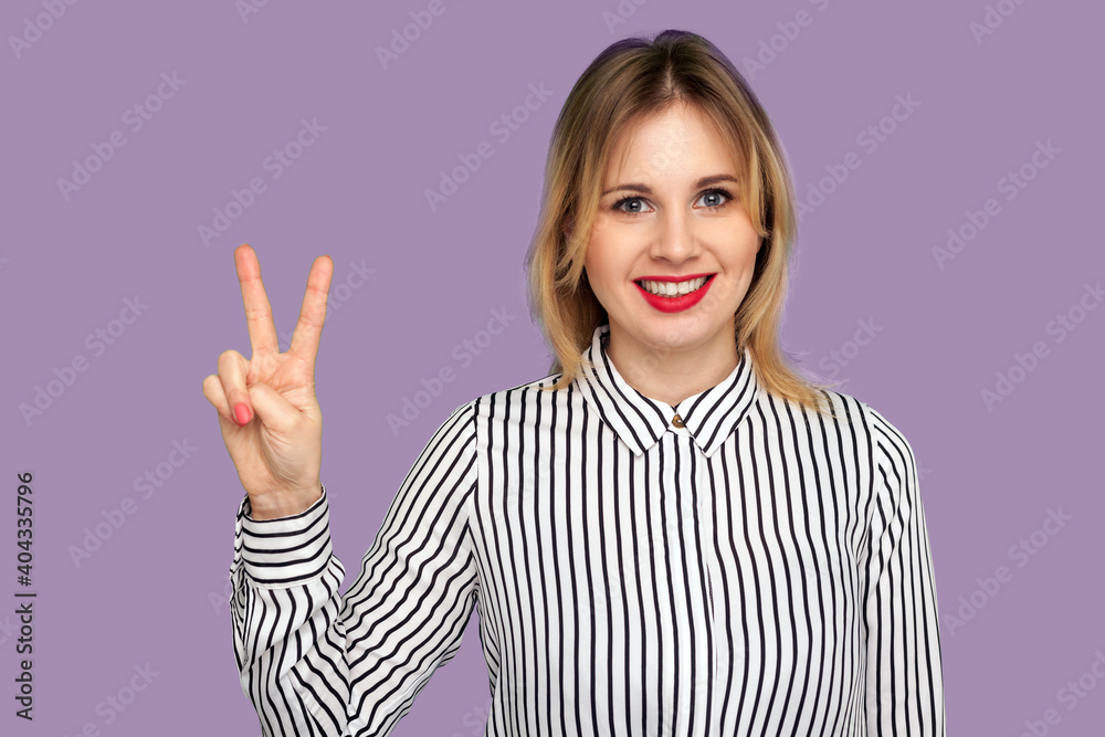 Portrait of happy attractive woman in striped blouse showing two finger sign, victory or peace gesture, looking at camera with charming toothy smile. indoor studio shot isolated on purple background Portrait of happy attractive woman in striped blouse showing two finger sign, victory or peace gesture, looking at camera with charming toothy smile. indoor studio shot isolated on purple background