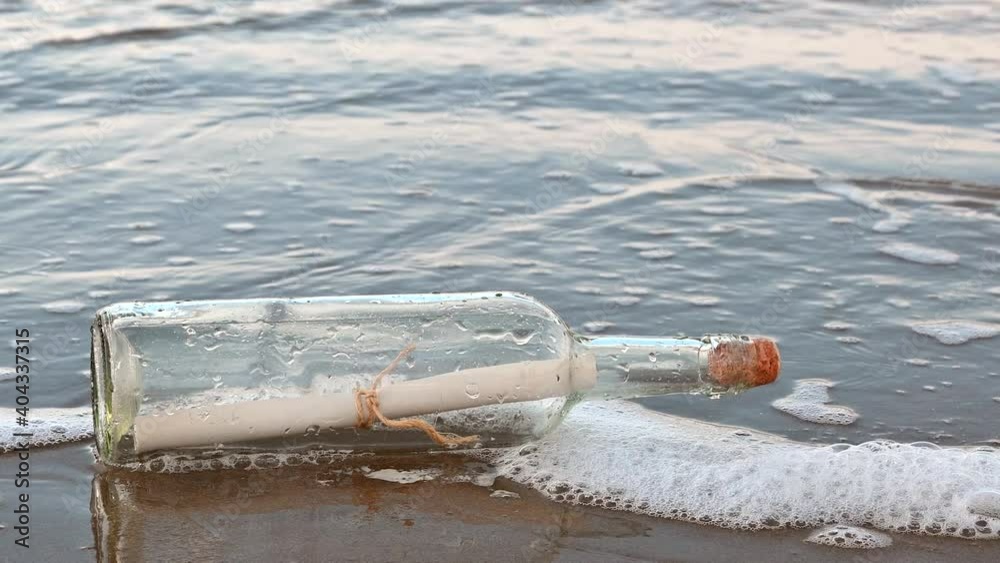 Closeup view of a message in a bottle lying on a wet sandy beach. Waves with foam come towards the bottle from the ocean. Bottle is horizontal with a cork in the opening.