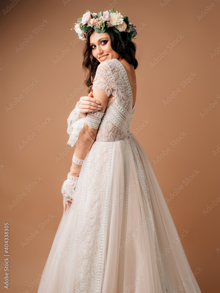 Happy bride in lush wedding dress posing in studio on brown background. Spring wedding dress with long lace sleeves and floral garland. Happy bride in lush wedding dress posing in studio on brown background. Spring wedding dress with long lace sleeves and floral garland.