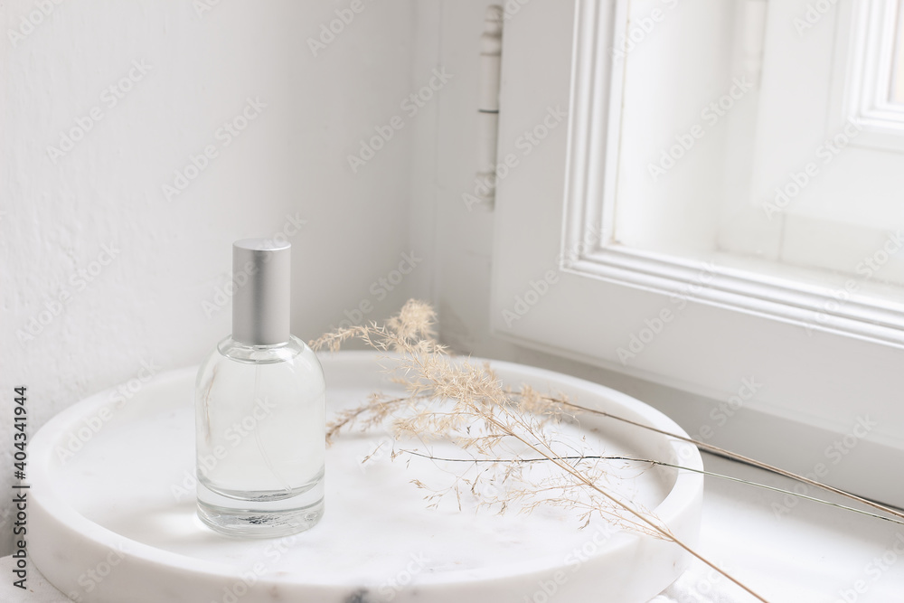 Feminine cosmetic still life scene. Glass bottle, flacon with perfume or eau de toilette and dry grass on white marble tray near window. Selective focus, blurred window sill background. Feminine cosmetic still life scene. Glass bottle, flacon with perfume or eau de toilette and dry grass on white marble tray near window. Selective focus, blurred window sill background.