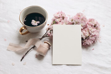 Wedding stationery mock-up. Blank greeting card and cup of coffee. White linen tablecloth background. Pink blossoming cherry tree branches and ribbon. Feminine still life composition. Flat lay,top.