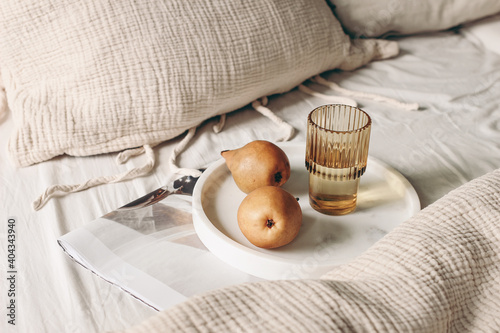 Autumn, summer breakfast in bed composition. Glass of water, folded newspapers and pear fruit on white marble tray. Champagne beige muslin cotton bed linen. Velvet cushions. Lifestyle, interior. photo