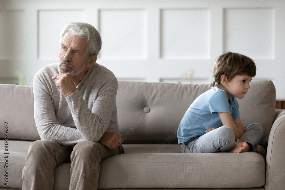 Upset senior grandfather and small 6s grandson sit back to back on couch avoid talking after fight. Unhappy elderly grandparent and little boy child have misunderstanding. Generation gap concept.