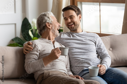 Overjoyed mature father and adult son sit relax on couch at home drink coffee or tea talking on weekend. Happy elderly dad and grownup man child have fun laugh enjoying family time together.