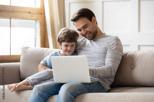 Smiling young Caucasian father and little son sit relax on sofa in living room use laptop together. Happy dad and small boy child rest on couch at home watch funny video on computer on family weekend.