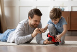 Caring happy young Caucasian father feel playful teach small 6s son repair fix toy car at home. Loving smiling dad lying on floor have fun play engaged in funny game activity with little boy child.