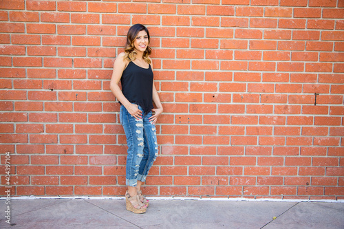 Hispanic young woman standing against a red brick wall wearing ripped jeans