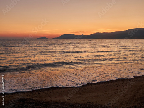 Sunset landscape with sea and mountains at the horizon. Sea coast in the dusk. Evening surf on sandy beach