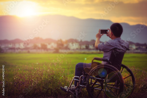 Close up of a paralyzed man sitting on wheelchair took a photo of the sunset by smartphone at rice green field with the blurred village and mountain background in the evening 