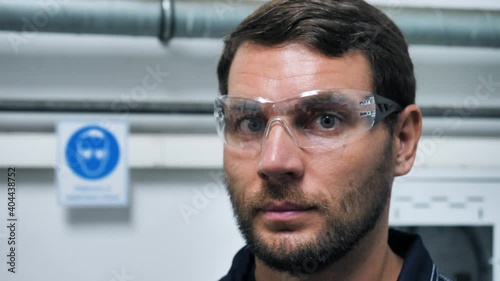 Close Up Portrait of Industry Engineer Worker In Manufacturing Factory Looks At Camera, Puts On Safety Glasses. Young Handsome Workman Mechanic Foreman In Protective Goggles In Industrial Plant.