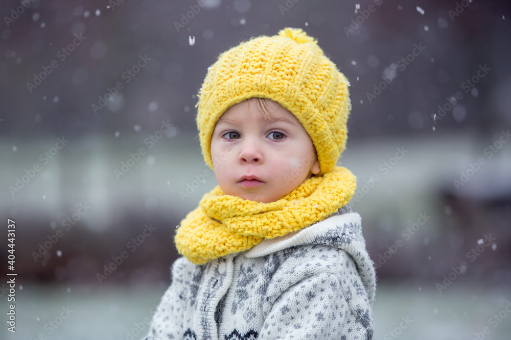 Beautiful blond toddler child, boy, with handmade knitted sweater playing in the park with first snow Beautiful blond toddler child, boy, with handmade knitted sweater playing in the park with first snow