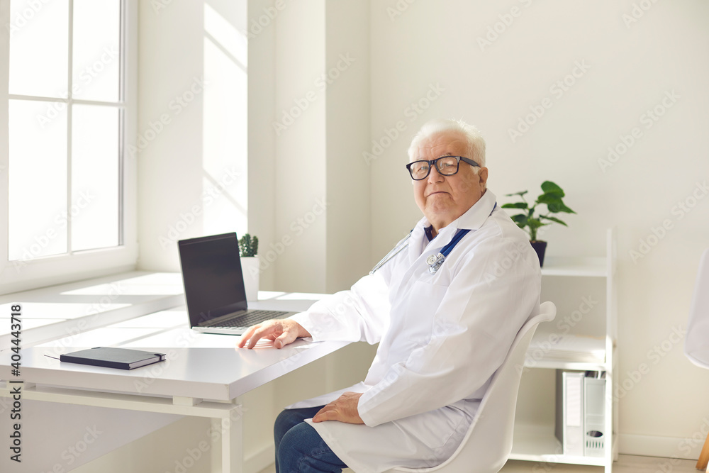 Positive senior professional doctor in uniform sitting with laptop in medical office and looking at camera