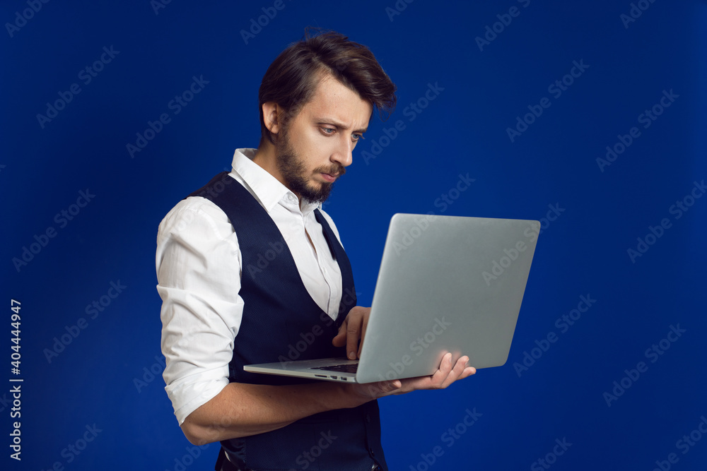 male businessman with a beard in a white shirt and vest holds a laptop on a blue background