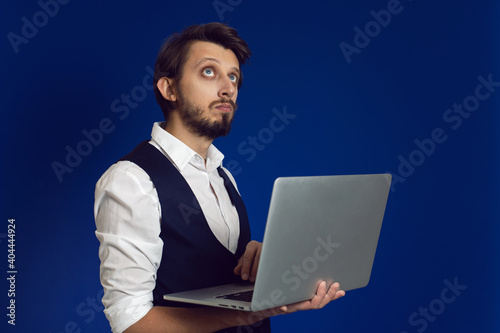 male businessman with a beard in a white shirt and vest holds a laptop on a blue background © saulich84