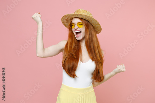 Young happy positive excited caucasian redhead woman 20s ginger long hair wear straw hat glasses summer clothes dancing clenching fists singing isolated on pastel pink background studio portrait.