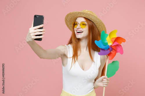 Young smiling funny redhead ginger woman 20s wear straw hat glasses summer clothes look camera holding toy windmill do selfie shot on mobile phone isolated on pastel pink background studio portrait.