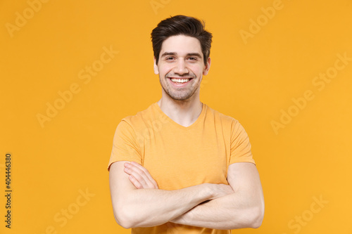Young smiling unshaved caucasian handsome man 20s years old in casual basic blank print design t-shirt look camera holding hands crossed folded isolated on yellow color background studio portrait.
