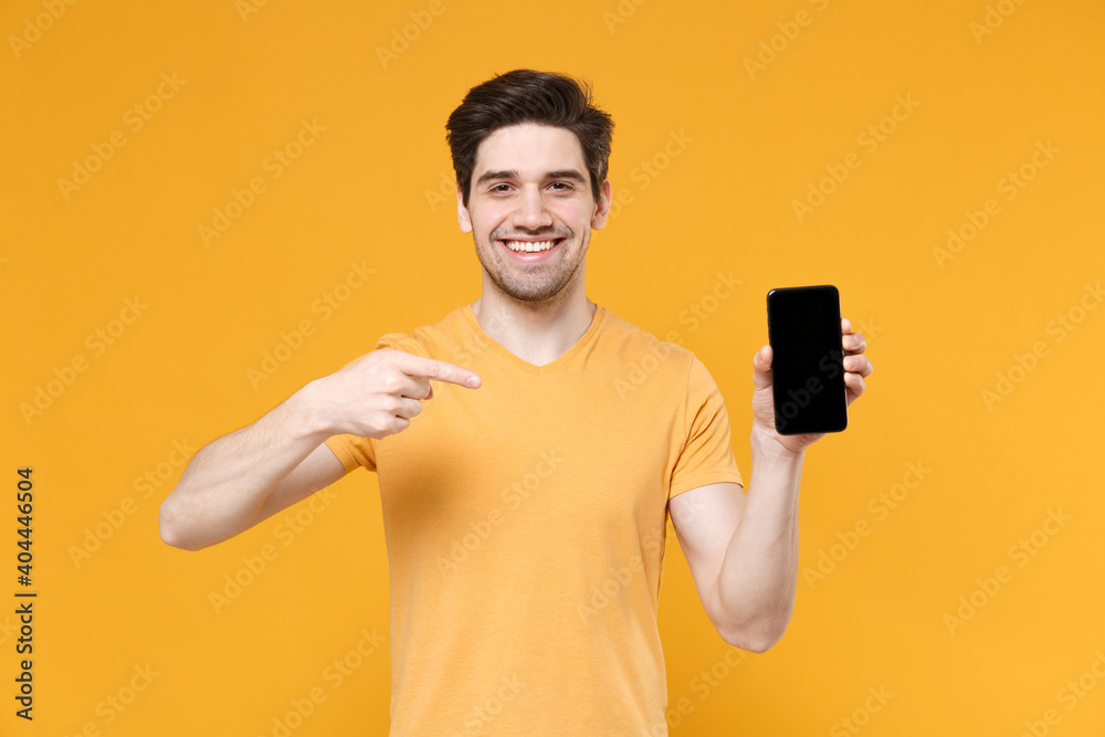 Young smiling man 20s in casual print design t-shirt hold point index finger on mobile cell phone with blank screen workspace copy space mock up isolated on yellow background studio portrait. Young smiling man 20s in casual print design t-shirt hold point index finger on mobile cell phone with blank screen workspace copy space mock up isolated on yellow background studio portrait.