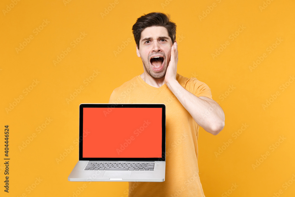 Young shocked confused troubled unshaved man 20s in basic t-shirt using laptop pc computer with blank screen workspace copy space mock up spreading hand isolated on yellow background studio portrait