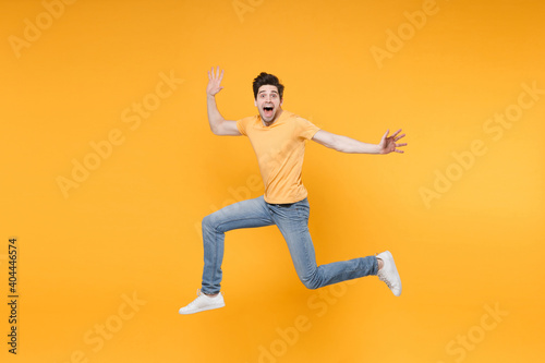 Full length side view of young overjoyed caucasian excited fun surprised man 20s wearing casual basic t-shirt jeans high jumping up spreading hands isolated on yellow color background studio portrait.