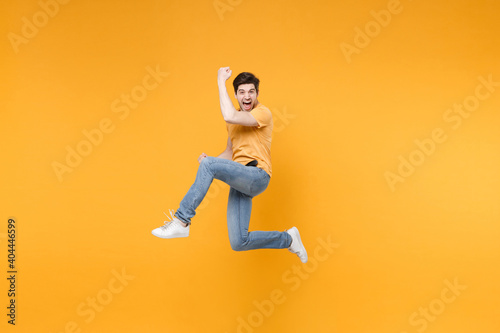 Full length side view of young overjoyed excited surprised man in casual t-shirt jeans high jump up do winner gesture with clenching fists look camera isolated on yellow background studio portrait.