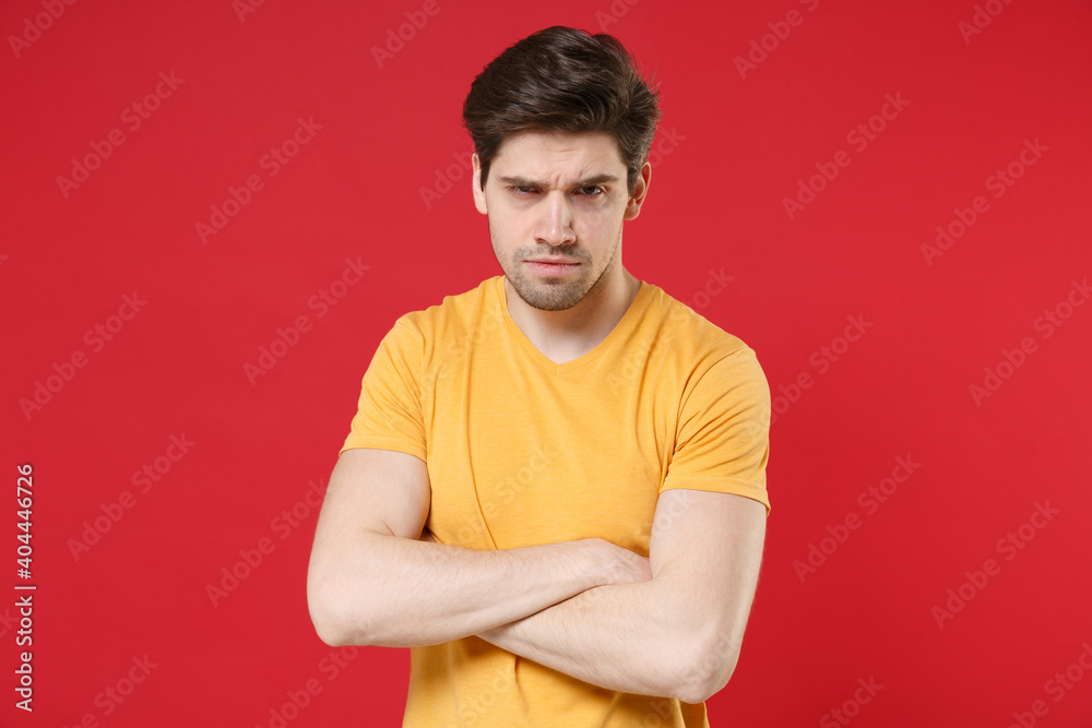 Young sceptical distrustful unshaved sad displeased caucasian man 20s in casual basic yellow t-shirt holding crossed hands folded looking camera isolated on red color background studio portrait Young sceptical distrustful unshaved sad displeased caucasian man 20s in casual basic yellow t-shirt holding crossed hands folded looking camera isolated on red color background studio portrait