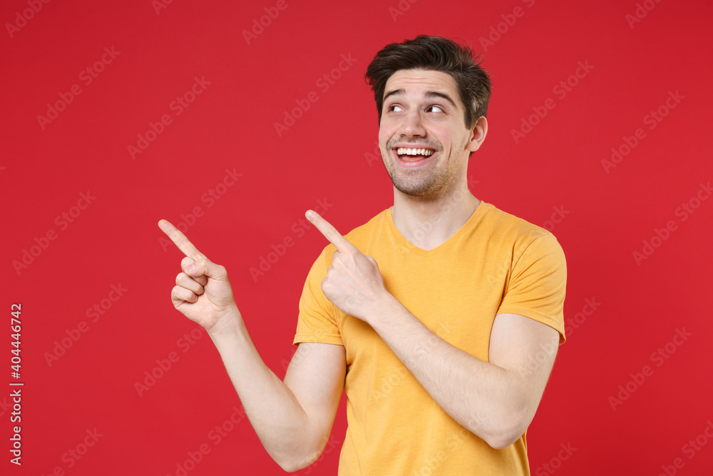 Young smiling unshaved cheerful caucasian man 20s in casual yellow t-shirt point index two fingers aside on workspace copy space area look aside isolated on red color background studio portrait. Young smiling unshaved cheerful caucasian man 20s in casual yellow t-shirt point index two fingers aside on workspace copy space area look aside isolated on red color background studio portrait.