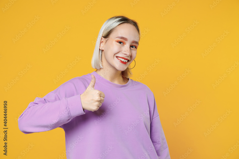 Smiling fun young blonde caucasian woman 20s bob haircut showing thumbs up wearing casual basic purple shirt looking camera charming toothy smile isolated on yellow color background studio portrait. Smiling fun young blonde caucasian woman 20s bob haircut showing thumbs up wearing casual basic purple shirt looking camera charming toothy smile isolated on yellow color background studio portrait.
