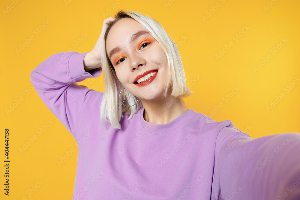 Smiling young blonde caucasian woman bob haircut bright makeup wearing basic purple shirt close up doing selfie shot on mobile phone open mouth isolated on yellow color background studio portrait. Smiling young blonde caucasian woman bob haircut bright makeup wearing basic purple shirt close up doing selfie shot on mobile phone open mouth isolated on yellow color background studio portrait.