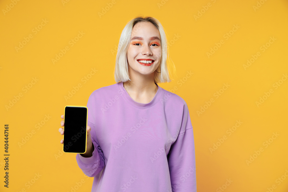 Young smiling blonde caucasian woman bob haircut in basic purple shirt holding mobile cell phone with blank screen workspace copy space mock up isolated on yellow color background studio portrait. Young smiling blonde caucasian woman bob haircut in basic purple shirt holding mobile cell phone with blank screen workspace copy space mock up isolated on yellow color background studio portrait.