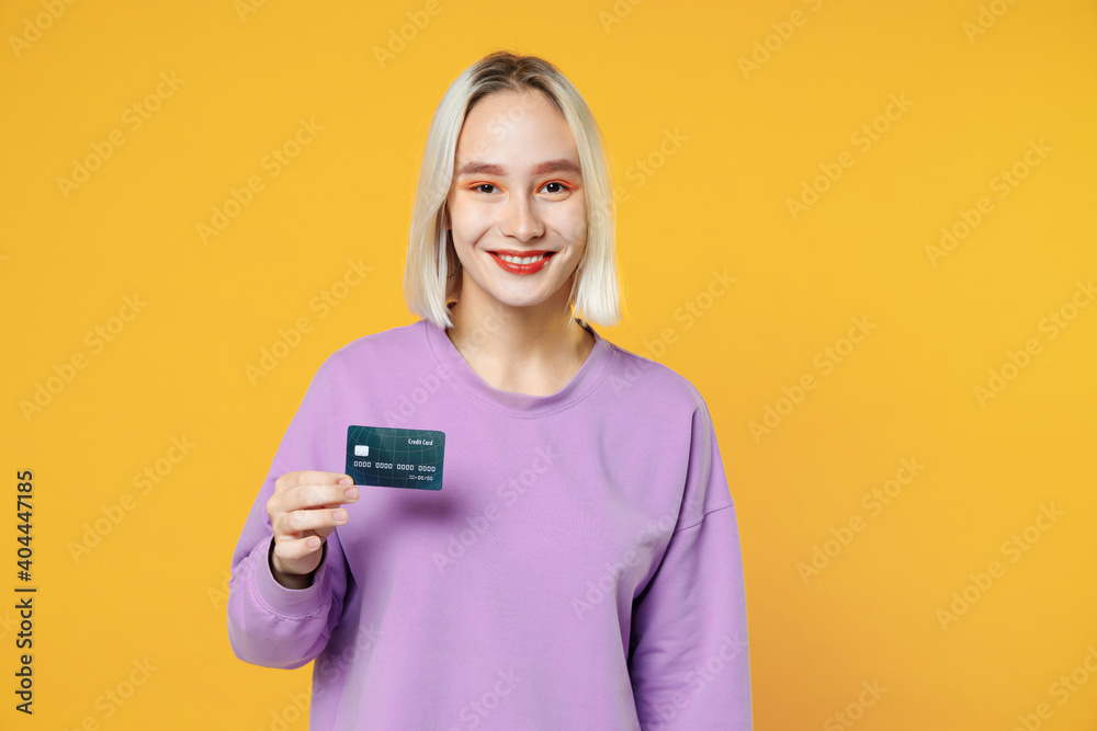 Smiling young blonde caucasian happy woman 20s with bob haircut bright makeup wearing casual basic purple shirt holding in hand credit bank card isolated on yellow color background studio portrait.