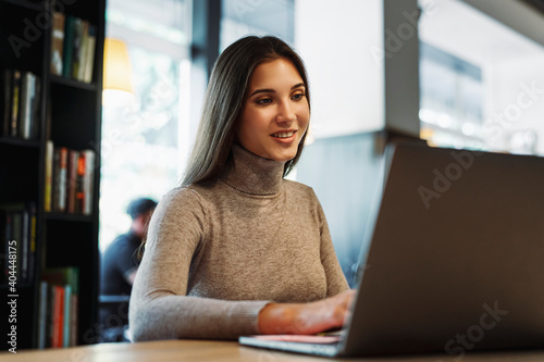 Attractive business woman sits at table in front of laptop, works remotely while in cafe. Freelancer discussing work using video communication. Beautiful brunette girl uses gadgets for work. 