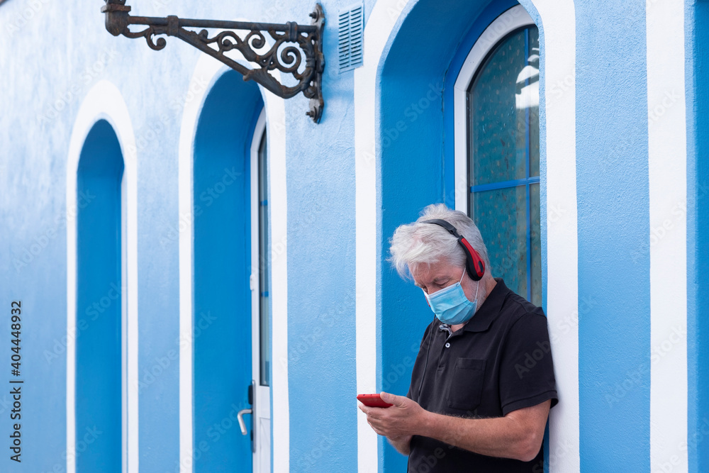 Senior man standing against a blue wall looking at mobile phone, listening to music with headphones. Wearing protective face mask due to coronavirus