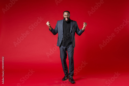 Full length of joyful young african american business man 20s wearing classic jacket suit standing doing winner gesture clenching fists isolated on bright red color wall background studio portrait. © ViDi Studio