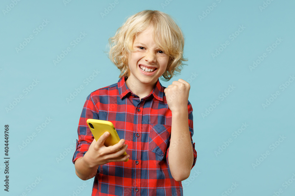 Happy little curly kid boy 10s in red checkered shirt using mobile cell phone typing sms message doing winner gesture isolated on blue background children studio portrait. Childhood lifestyle concept. Happy little curly kid boy 10s in red checkered shirt using mobile cell phone typing sms message doing winner gesture isolated on blue background children studio portrait. Childhood lifestyle concept.