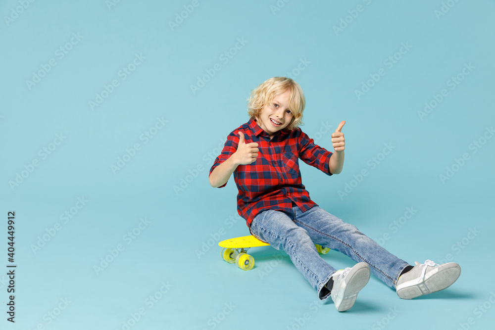 Full length of funny little curly kid boy 10s in basic red checkered shirt sitting on skateboard showing thumbs up isolated on blue background children studio portrait. Childhood lifestyle concept. Full length of funny little curly kid boy 10s in basic red checkered shirt sitting on skateboard showing thumbs up isolated on blue background children studio portrait. Childhood lifestyle concept.
