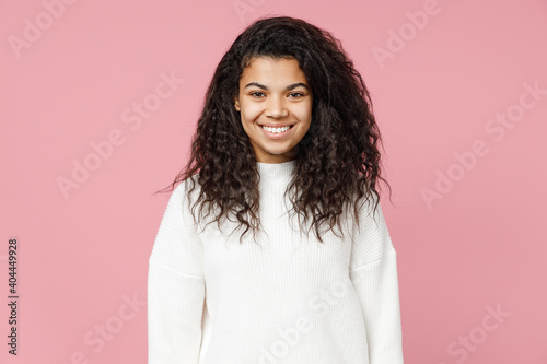 Young smiling happy cute attractive african american woman 20s curly hair wear white casual knitted sweater looking camera isolated on pastel pink background studio portrait People lifestyle concept.
