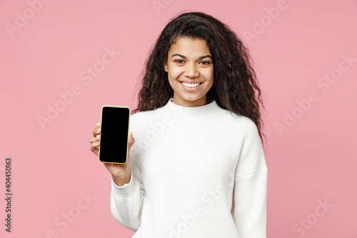 Young smiling attractive african american curly woman 20s wear white casual knitted sweater hold mobile cell phone with blank screen workspace area isolated on pastel pink background studio portrait.