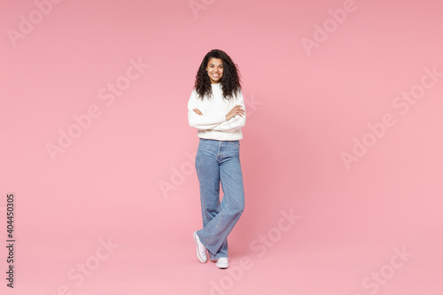 Full length young smiling african american woman 20s curly hair wear white casual knitted sweater jeans looking camera hold hands crossed folded isolated on pastel pink background studio portrait.