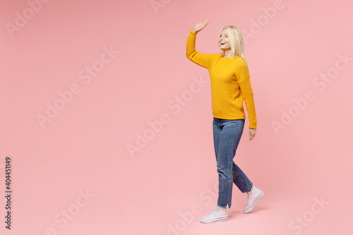 Full length side view of funny elderly gray-haired blonde woman lady 40s 50s years old in yellow sweater waving and greeting with hand as notices someone isolated on pink background studio portrait.