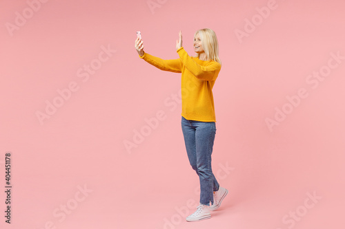 Full length side view of funny elderly gray-haired blonde woman lady 40s 50s in yellow sweater doing selfie shot on mobile phone greeting with hand isolated on pink color background studio portrait. photo