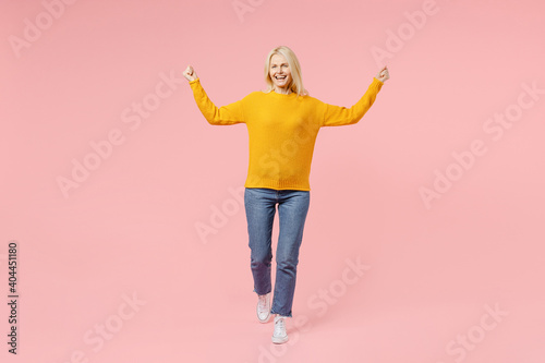Full length of happy elderly gray-haired blonde woman lady 40s 50s years old in yellow basic sweater standing doing winner gesture clenching fists isolated on pastel pink background studio portrait.