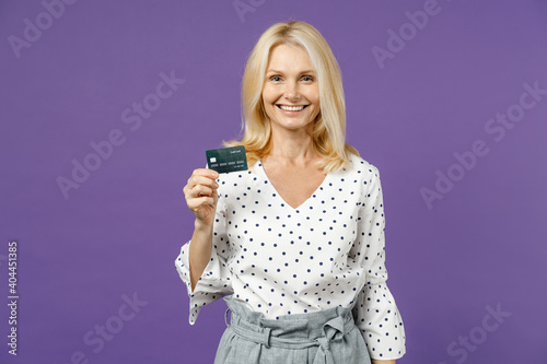 Smiling elderly gray-haired blonde woman lady 40s 50s years old in white dotted blouse standing hold in hand credit bank card looking camera isolated on bright violet color background studio portrait.