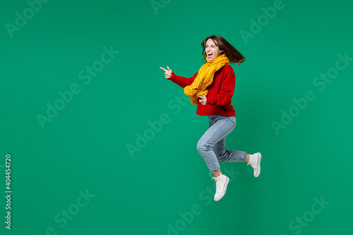 Full length side view of laughing cheerful young brunette woman 20s in casual knitted red sweater yellow scarf jumping pointing index fingers aside isolated on bright green background studio portrait.