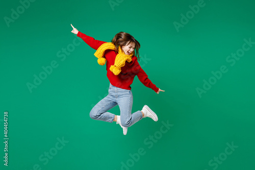 Full length side view of laughing funny young brunette woman 20s in casual knitted red sweater yellow scarf jumping pointing index fingers aside isolated on bright green background studio portrait.